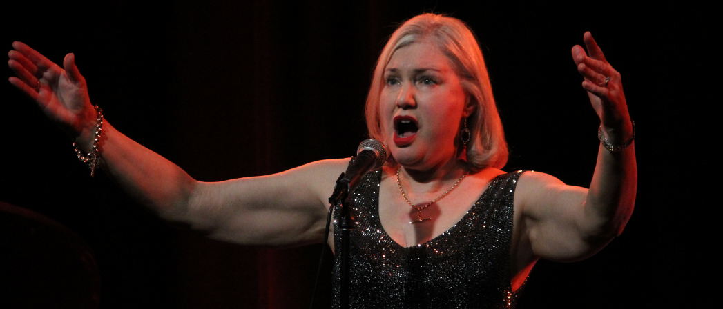 A blonde woman wearing a sparkly dress sings into a microphone while lifting both arms wide.